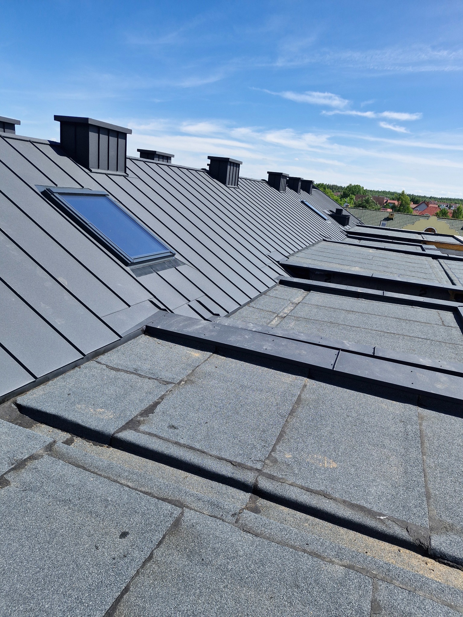 Szary dach z blachy trapezowej z oknem dachowym i kominami, sąsiadujący z dachem pokrytym papą na tle błękitnego nieba.