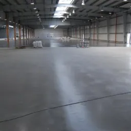 Gładka, szara powierzchnia w hali przemysłowej z widocznymi workami materiałów budowlanych i pojemnikami w tle. Oświetlenie górne odbija się od świeżo wylanej posadzki.