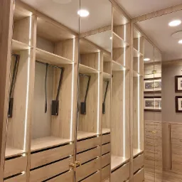 Wnętrze szafy wnękowej z jasnego drewna z oświetleniem LED, lustrzane drzwi przesuwne, widoczne półki, szuflady i pantografy na ubrania.
