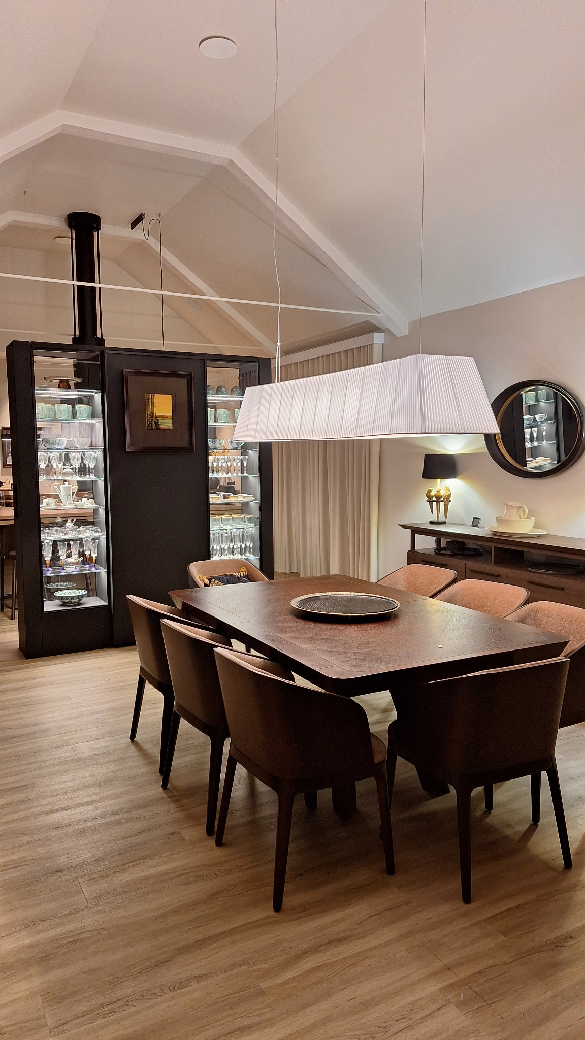 Jadalnia w nowoczesnym stylu z drewnianym stołem, krzesłami i oświetleniem wiszącym, z przeszkloną witryną z podświetleniem eksponującą szkło i ceramikę.