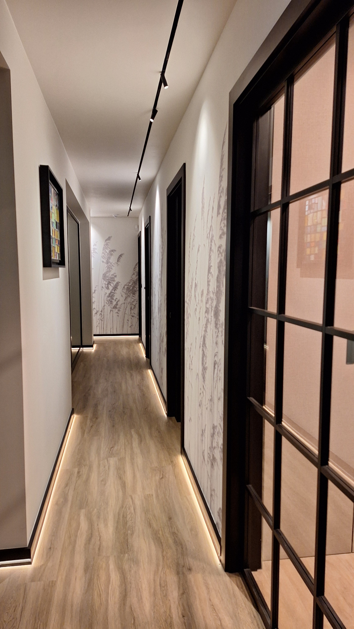 Nowoczesny korytarz z oświetleniem LED wzdłuż podłogi i na suficie, ściany ozdobione tapetą z motywem roślinnym, czarne ramy drzwi i okien.