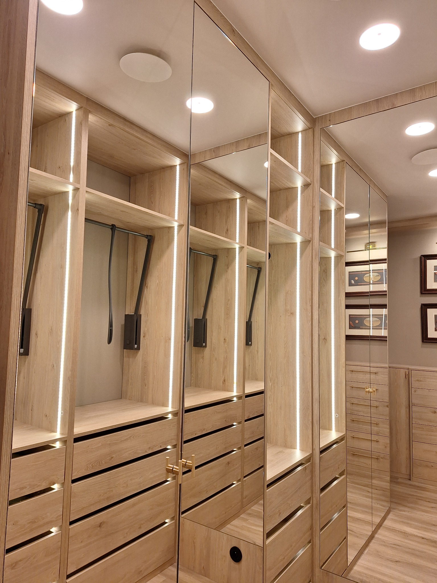 Wnętrze szafy wnękowej z jasnego drewna z oświetleniem LED, lustrzane drzwi przesuwne, widoczne półki, szuflady i pantografy na ubrania.