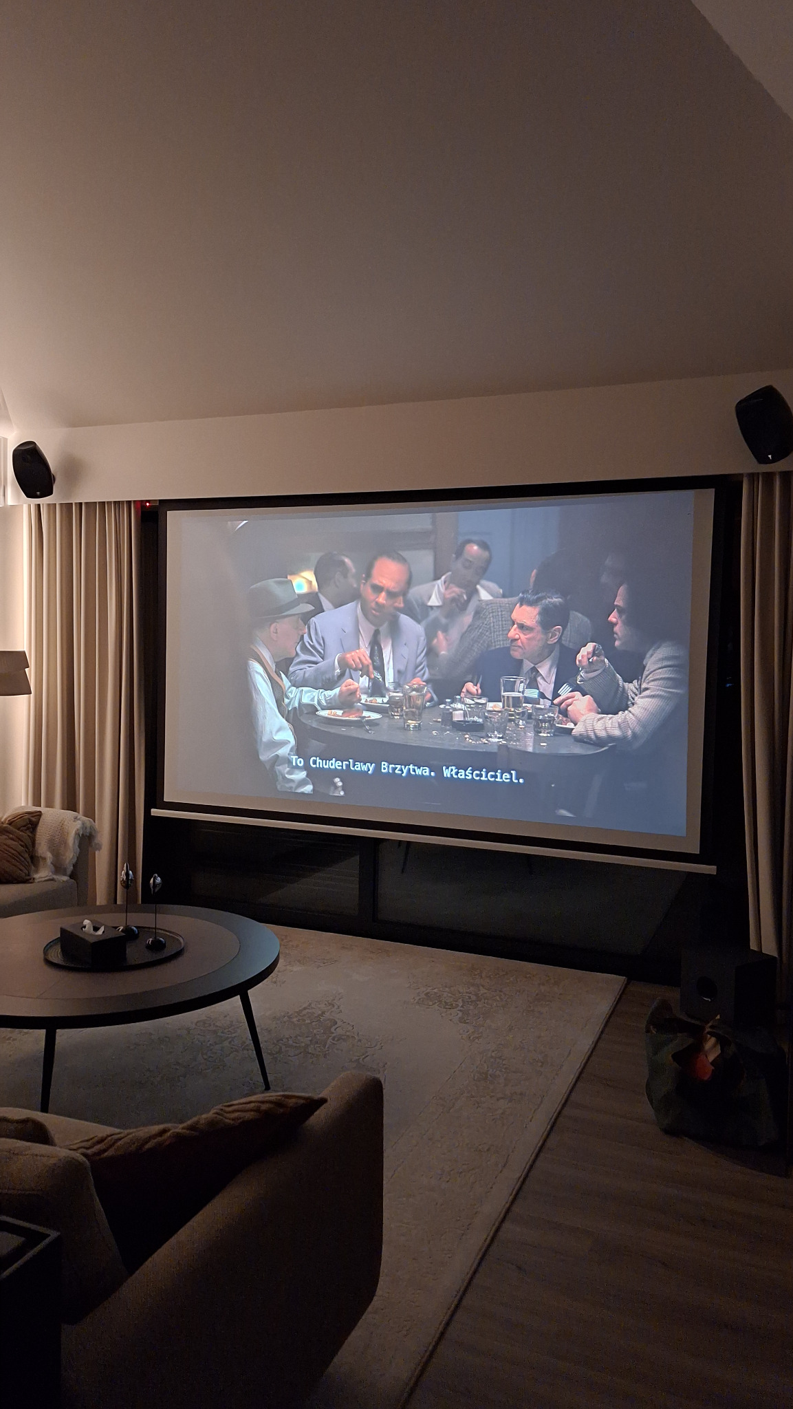 Salon z dużym ekranem projekcyjnym wyświetlającym scenę z filmu, głośniki na ścianie, zasłony i stolik kawowy w tle.