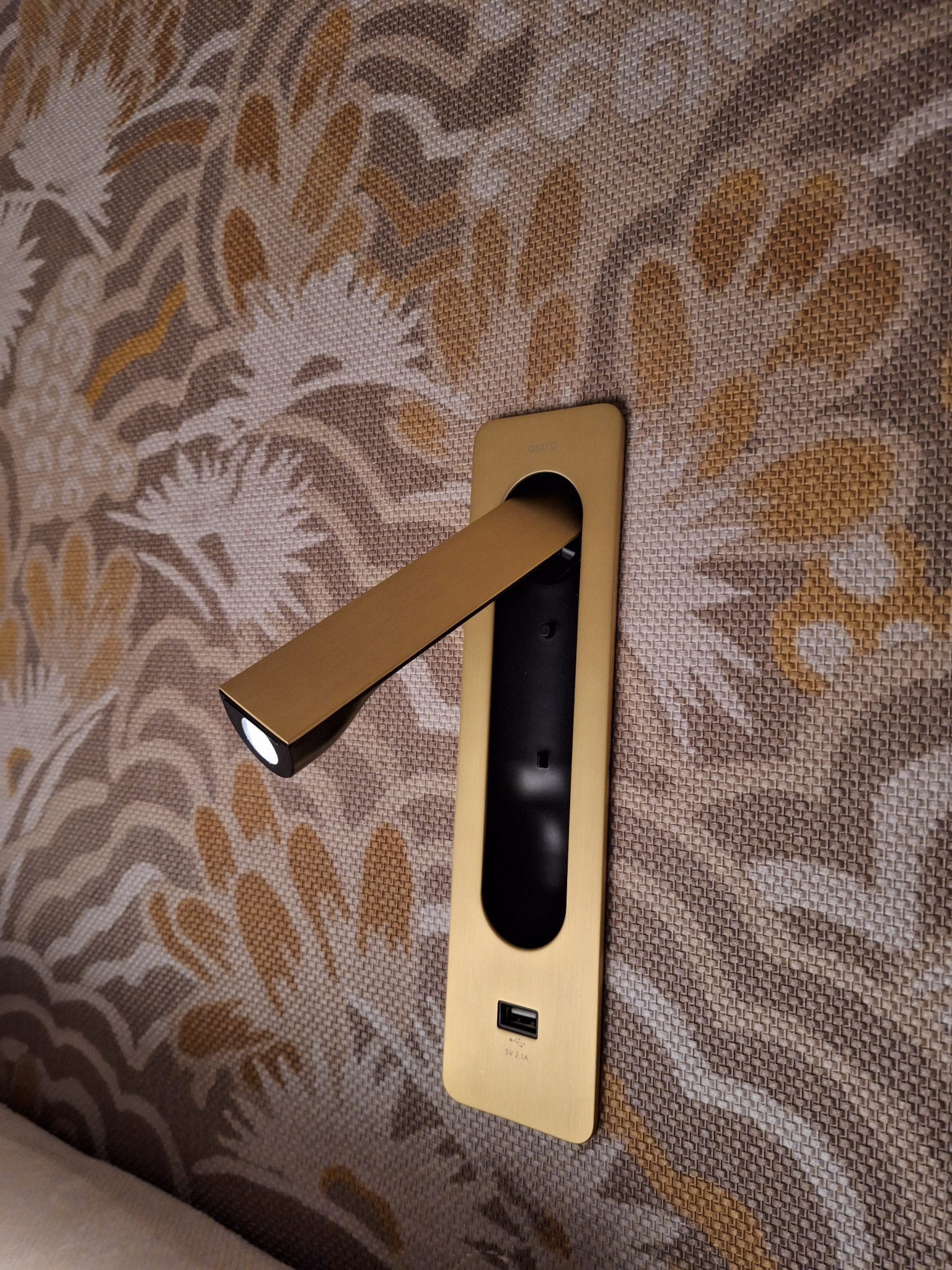 Złota lampka nocna LED z portem USB zamontowana na wzorzystej ścianie.