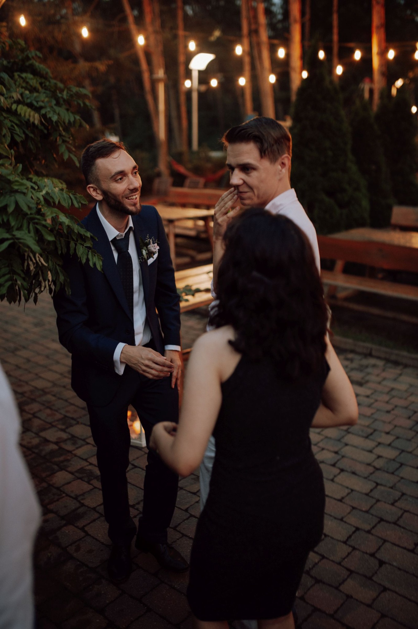 Uśmiechnięty mężczyzna w granatowym garniturze z butonierką rozmawia z dwoma osobami na zewnątrz, w tle dekoracyjne żarówki i drzewa.