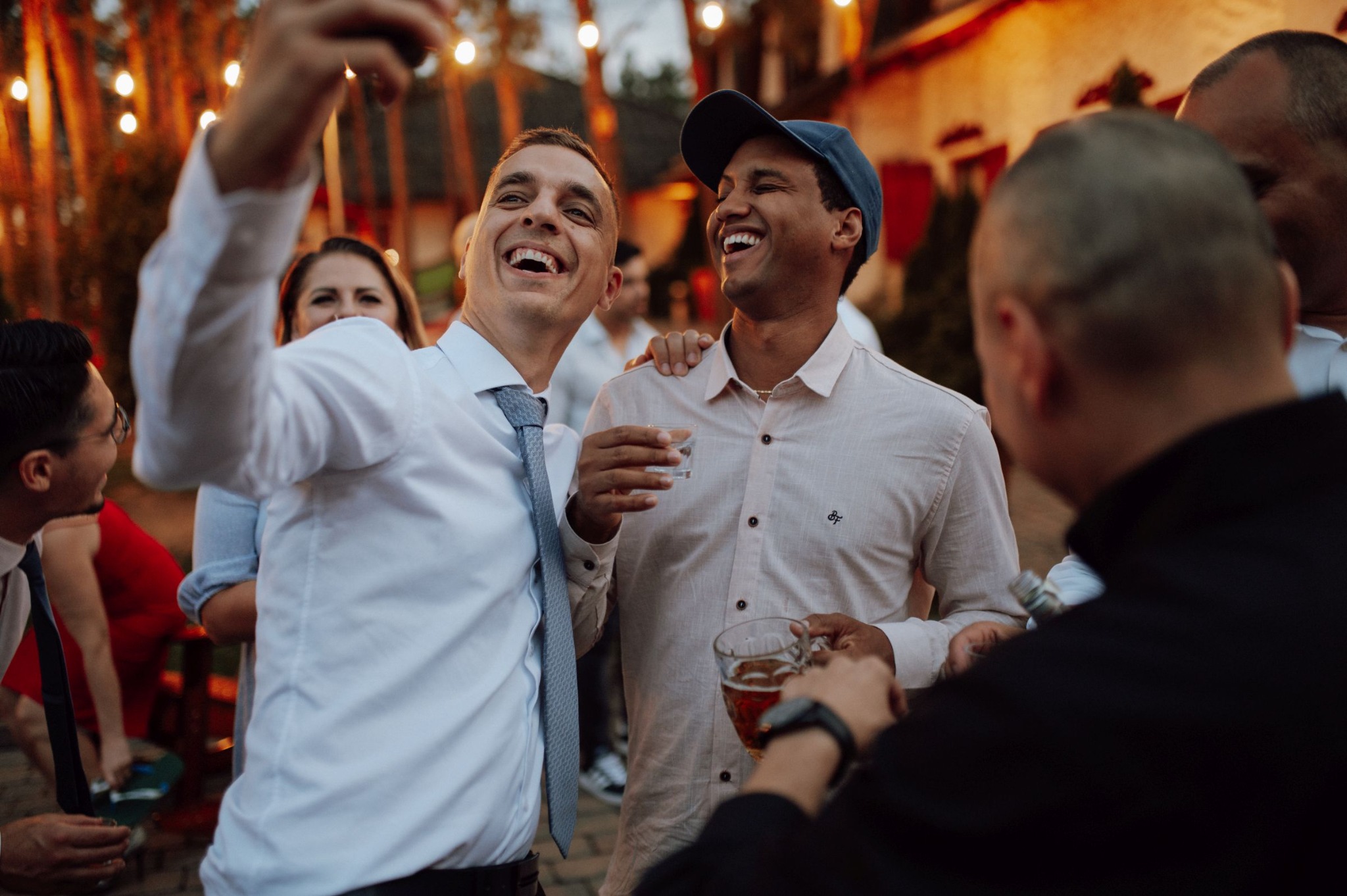 Radosny toast dwóch mężczyzn podczas przyjęcia plenerowego, jeden robi selfie, w tle rozświetlone girlandy żarówek.