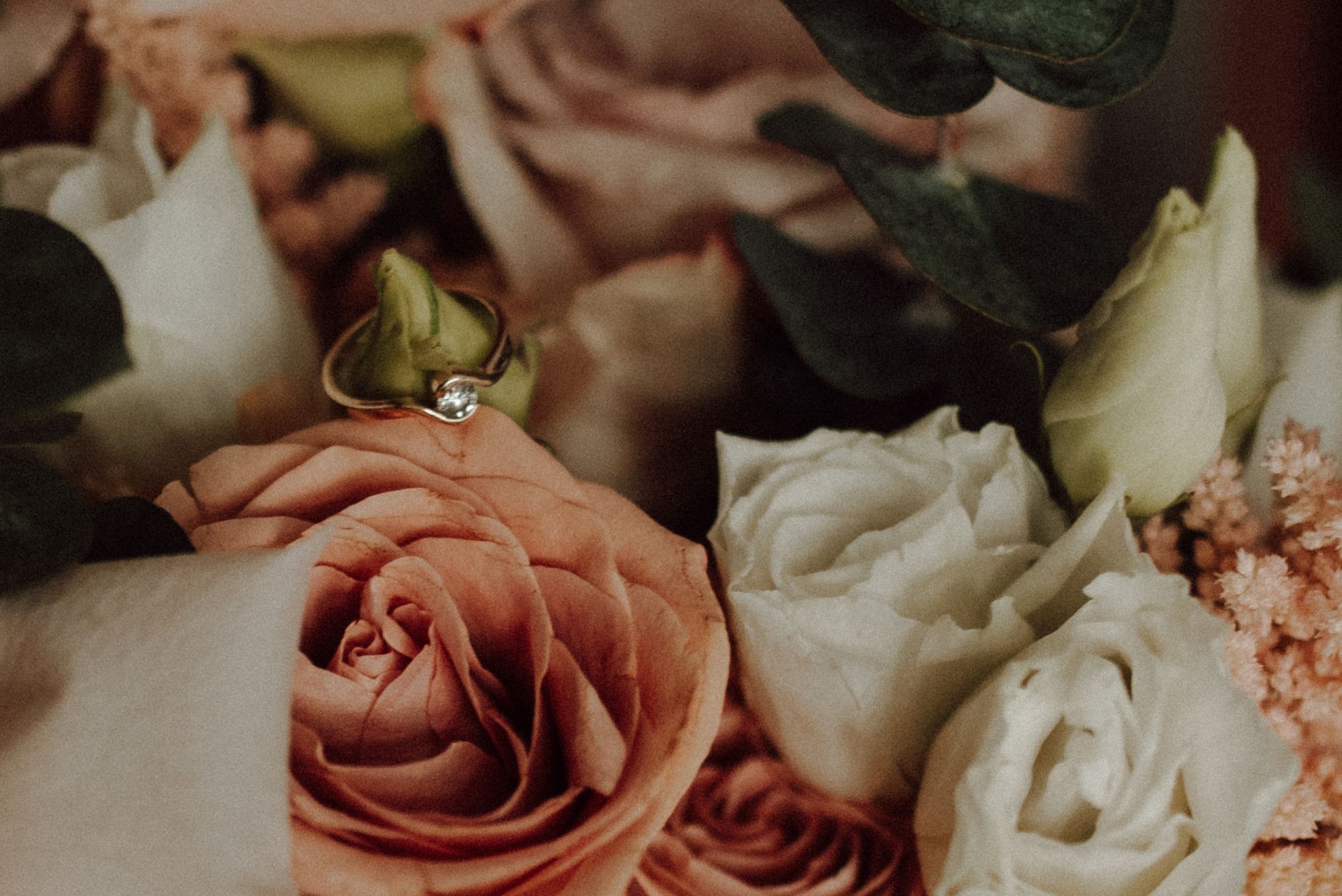 Złoty pierścionek zaręczynowy z pojedynczym diamentem spoczywa na brzoskwiniowej róży w bukiecie ślubnym z białych róż i zielonych liści eukaliptusa.