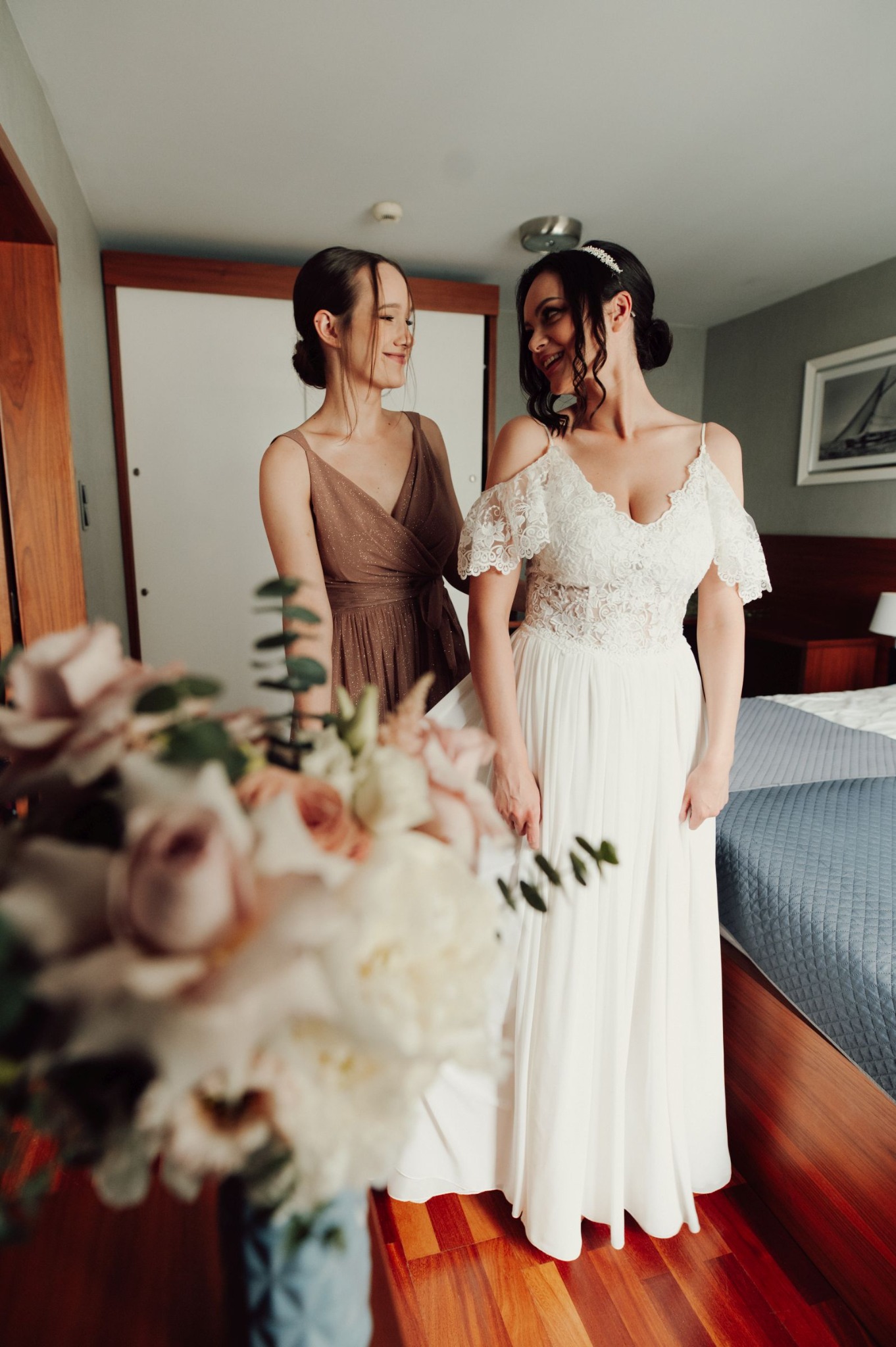Panna młoda w koronkowej sukni z odkrytymi ramionami, uśmiechnięta do druhny w sukience w kolorze taupe, bukiet kwiatów w pierwszym planie, przygotowania ślubne w pokoju hotelowym.