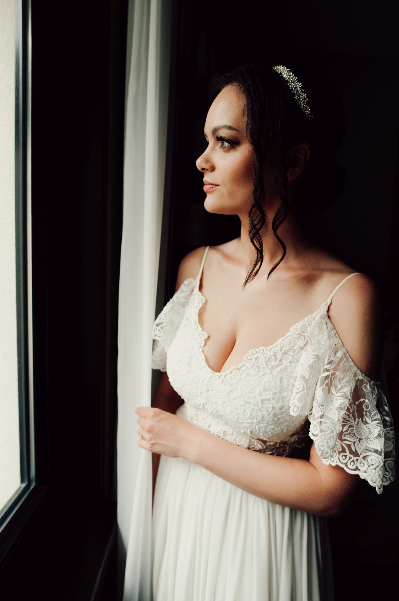 Panna młoda w koronkowej sukni z odkrytymi ramionami, z tiarą we włosach, spogląda przez okno trzymając w dłoni zwiewną zasłonę.