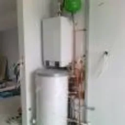 Biały bojler i piec zawieszone na ścianie, połączone miedzianymi rurami. Widoczny zielony element instalacji. Instalacja w pomieszczeniu z białymi ścianami.