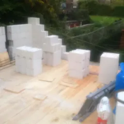 Budowa domu z bloczków betonu komórkowego na drewnianym szalunku, widoczne ułożone bloczki i drabina, w tle zieleń.