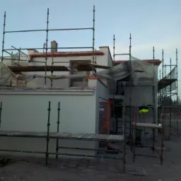 Dwukondygnacyjny dom w trakcie prac elewacyjnych, otoczony rusztowaniami i częściowo zakryty folią ochronną, widoczny fragment otynkowanej ściany i komin.