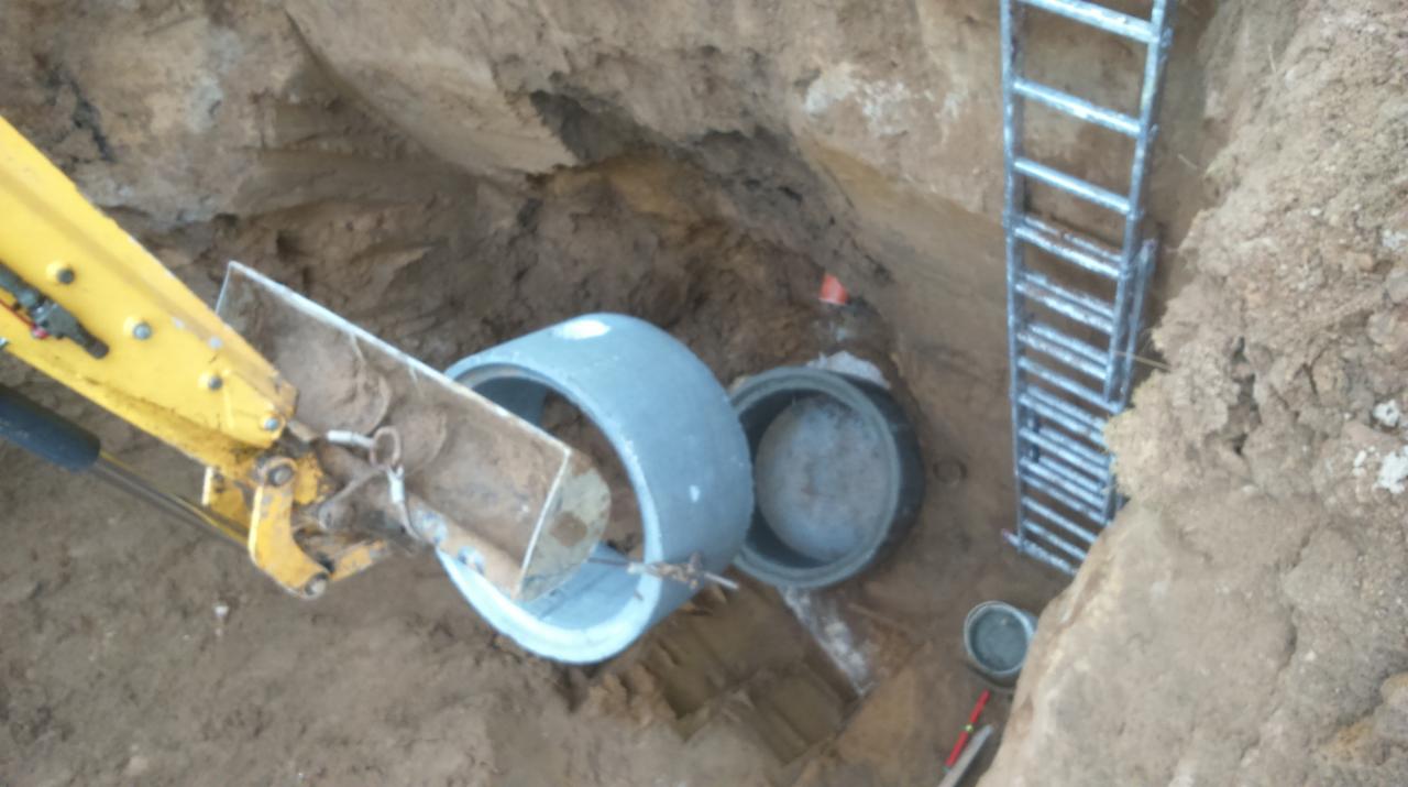 Żółta koparka umieszcza betonowy krąg w wykopie, obok stoi drabina, widoczne rury i poziomica.