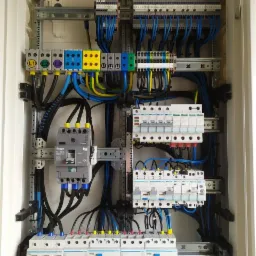 Rozdzielnica elektryczna z widocznymi bezpiecznikami, przewodami w kolorach niebieskim, żółtym i czarnym, zamontowana w białej obudowie, z szynami DIN i elementami oznaczonymi L1, L2, L3.