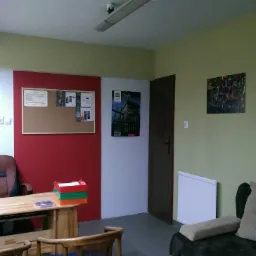 Wnętrze biura z biurkiem, komputerem, krzesłem obrotowym, tablicą korkową, kalendarzem ściennym i sofą. Na ścianach wiszą obrazy. Na biurku leżą kolorowe teczki.