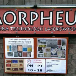 Szyld reklamowy firmy Morpheus z napisem 'Sztuka Technologii Laserowych' wiszący nad tablicami ogłoszeń z ofertą usług, godzinami otwarcia oraz informacją o przyjmowaniu zamówień na Pierwszą...