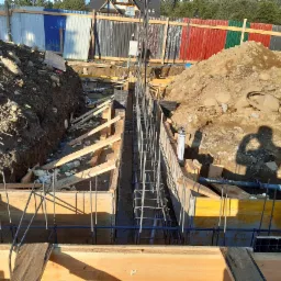 Fundamenty domu jednorodzinnego w trakcie budowy, z widocznym zbrojeniem i szalunkiem drewnianym, otoczone wykopaną ziemią i kolorowym ogrodzeniem z blachy falistej w tle.