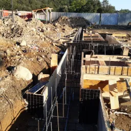 Budowa fundamentów: zbrojenie w szalunkach na gruncie z wykopaną ziemią i kamieniami, widoczne zabezpieczenia i narzędzia budowlane.