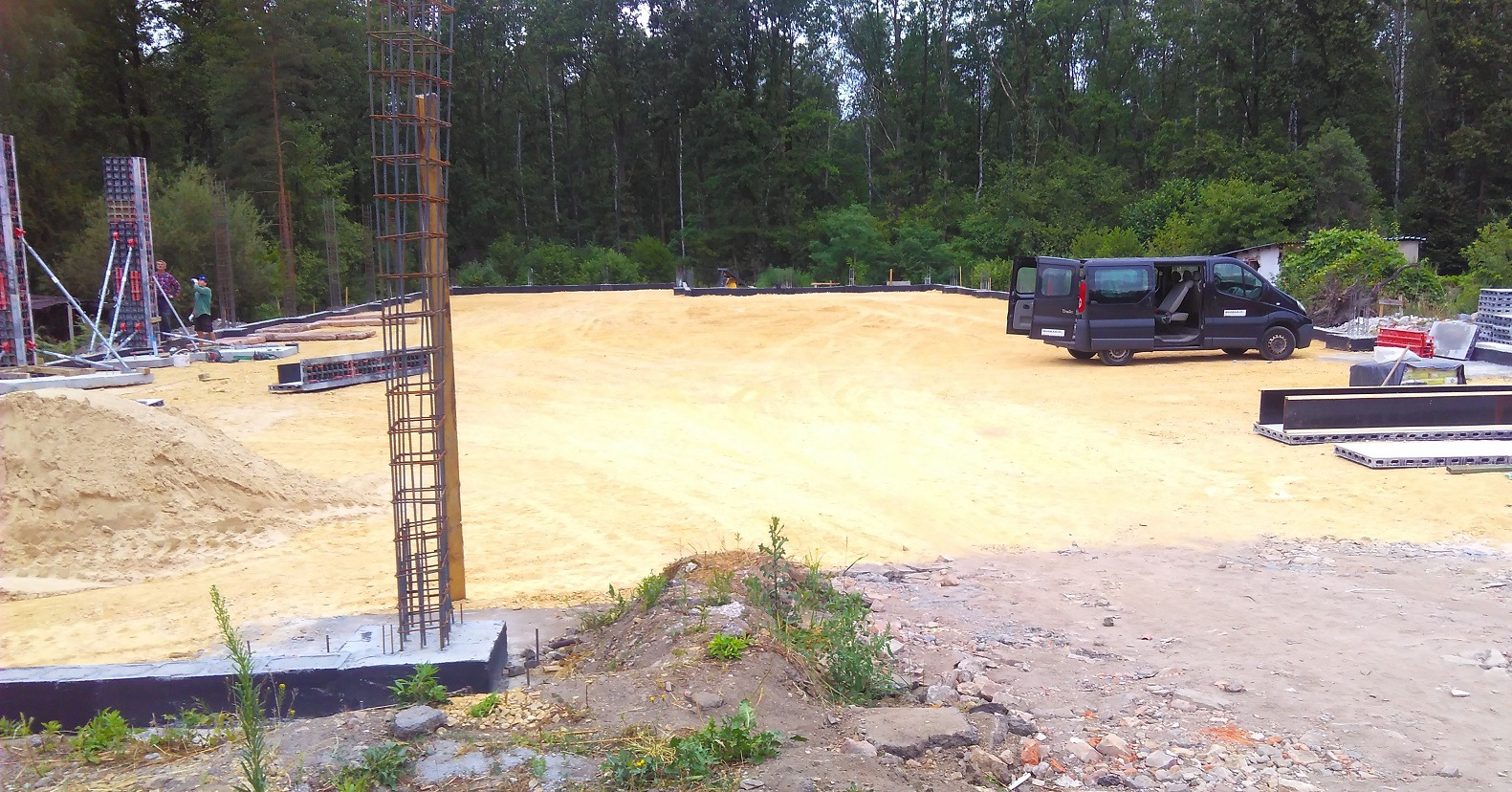 Przygotowany teren pod budowę z wystającymi zbrojeniami fundamentów, zaparkowany czarny van z otwartymi drzwiami, widoczne szalunki i piasek.