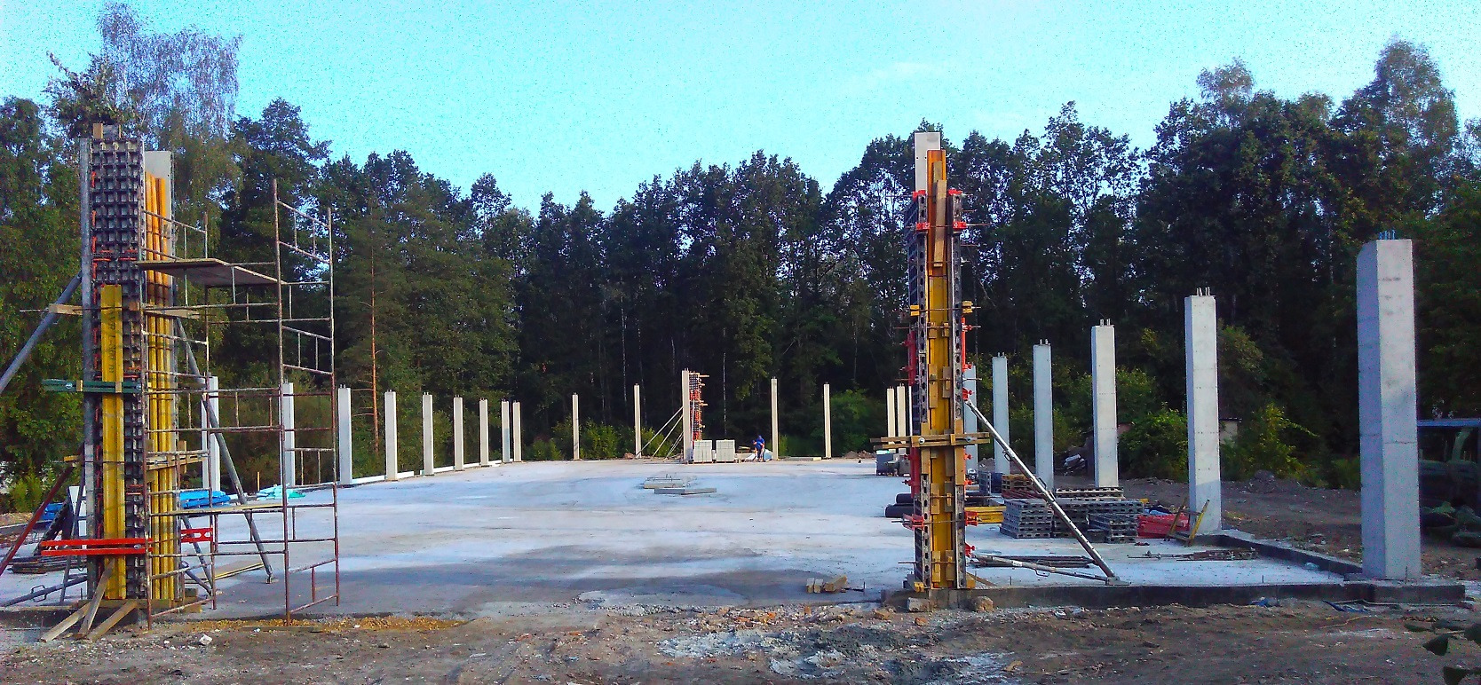 Wylewanie betonowych słupów konstrukcyjnych na placu budowy z użyciem szalunków systemowych i rusztowań, na tle lasu.