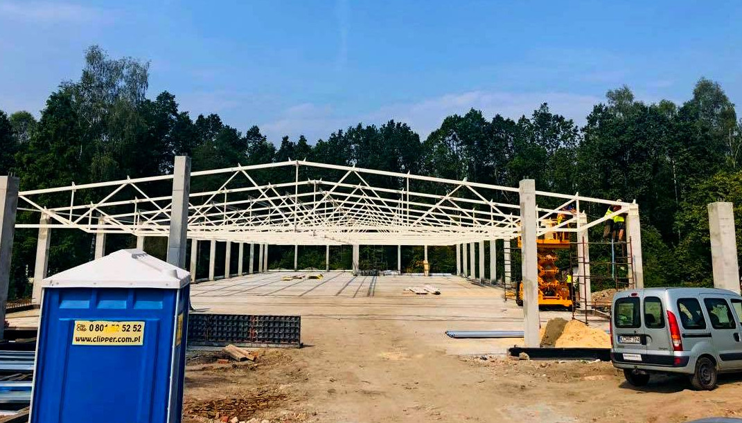 Wznoszenie konstrukcji stalowej hali na fundamencie betonowym, widoczny szkielet z białych belek, pracownik na podnośniku, przenośna toaleta na pierwszym planie.