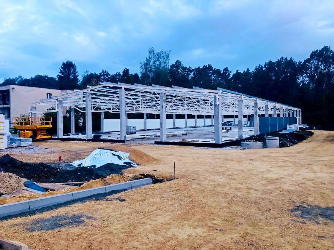 Budowa hali: stalowa konstrukcja z białymi słupami i dachem, widoczny podnośnik koszowy, teren budowy w trakcie prac ziemnych.
