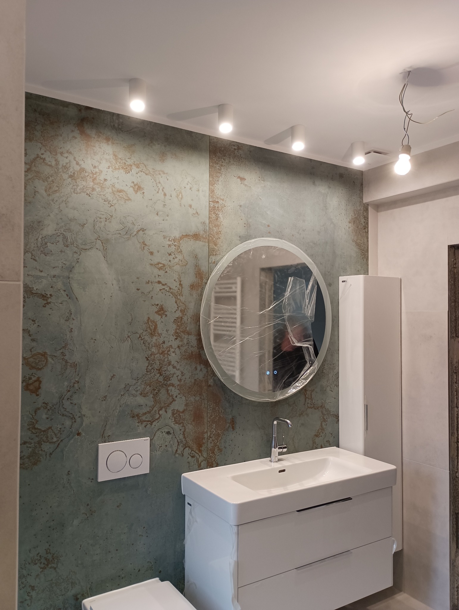 Nowoczesna łazienka z umywalką na szafce, okrągłym lustrem w folii ochronnej i ścianą z efektem patyny, oświetlona punktowymi lampami sufitowymi.