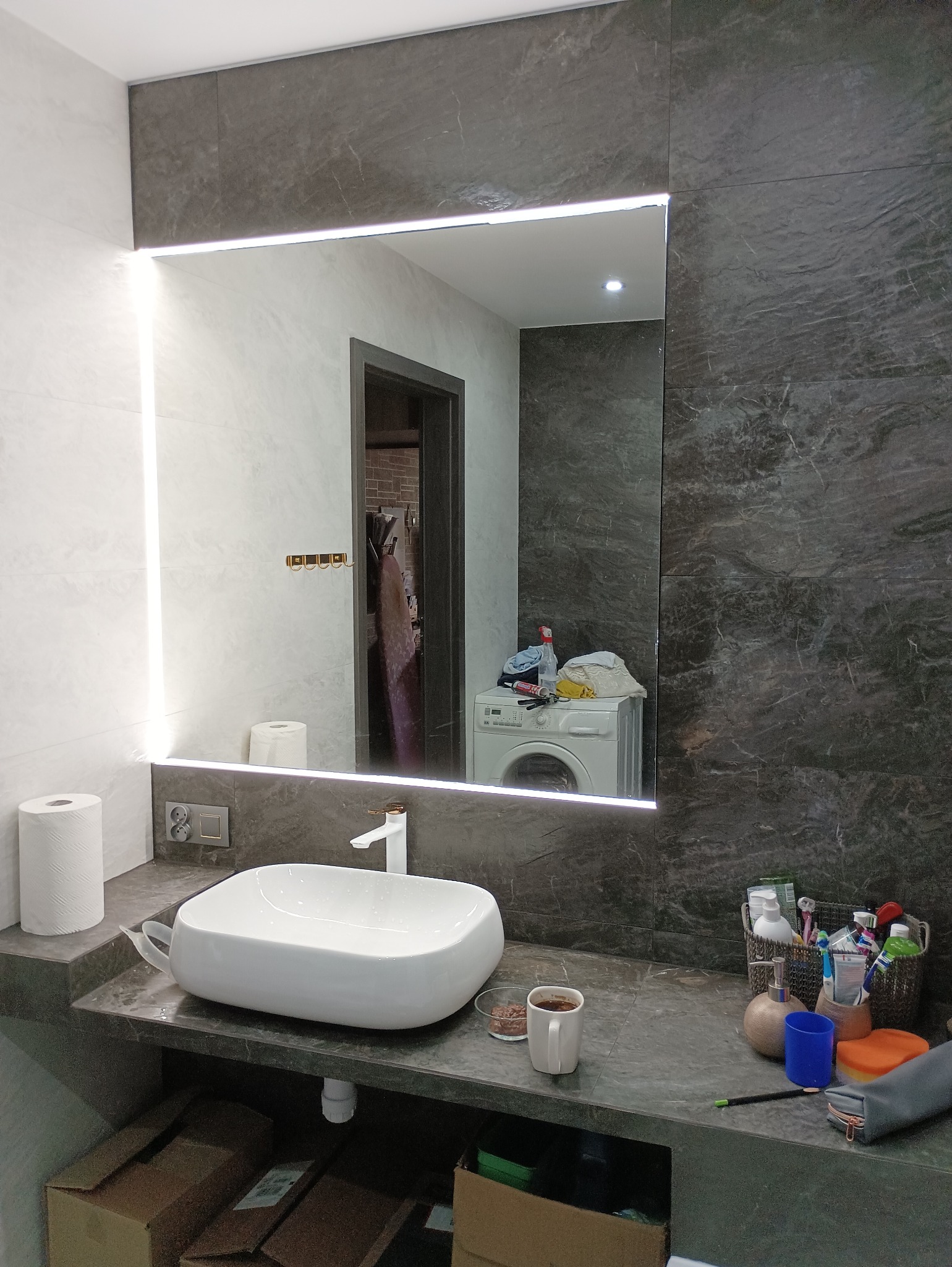 Nowoczesna łazienka z umywalką nablatową, podświetlanym lustrem, szarą marmurową okładziną ścian i pralką widoczną w odbiciu lustra.