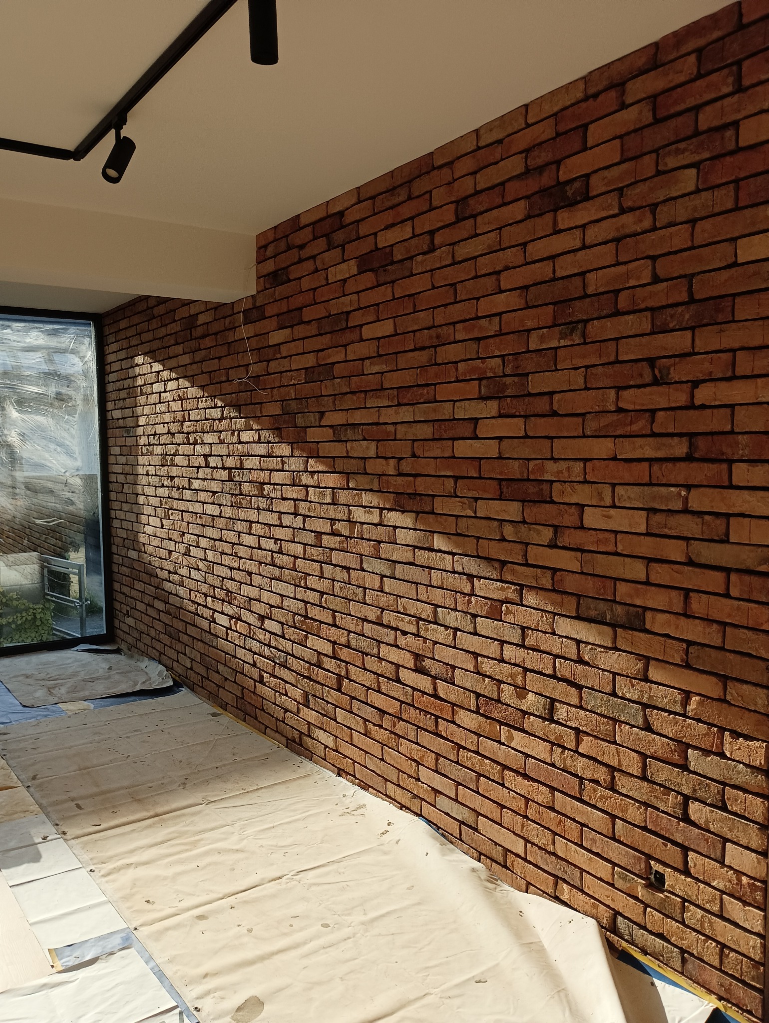 Ściana wewnętrzna wyłożona dekoracyjną czerwoną cegłą, oświetlona punktowo, z podłogą zabezpieczoną folią malarską.