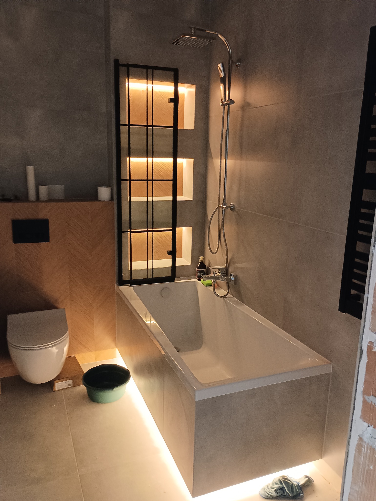Nowoczesna łazienka z podświetlaną wanną i półkami wnękowymi, szara płytka imitująca beton, drewniane akcenty na zabudowie toalety, bateria wannowa z deszczownicą.