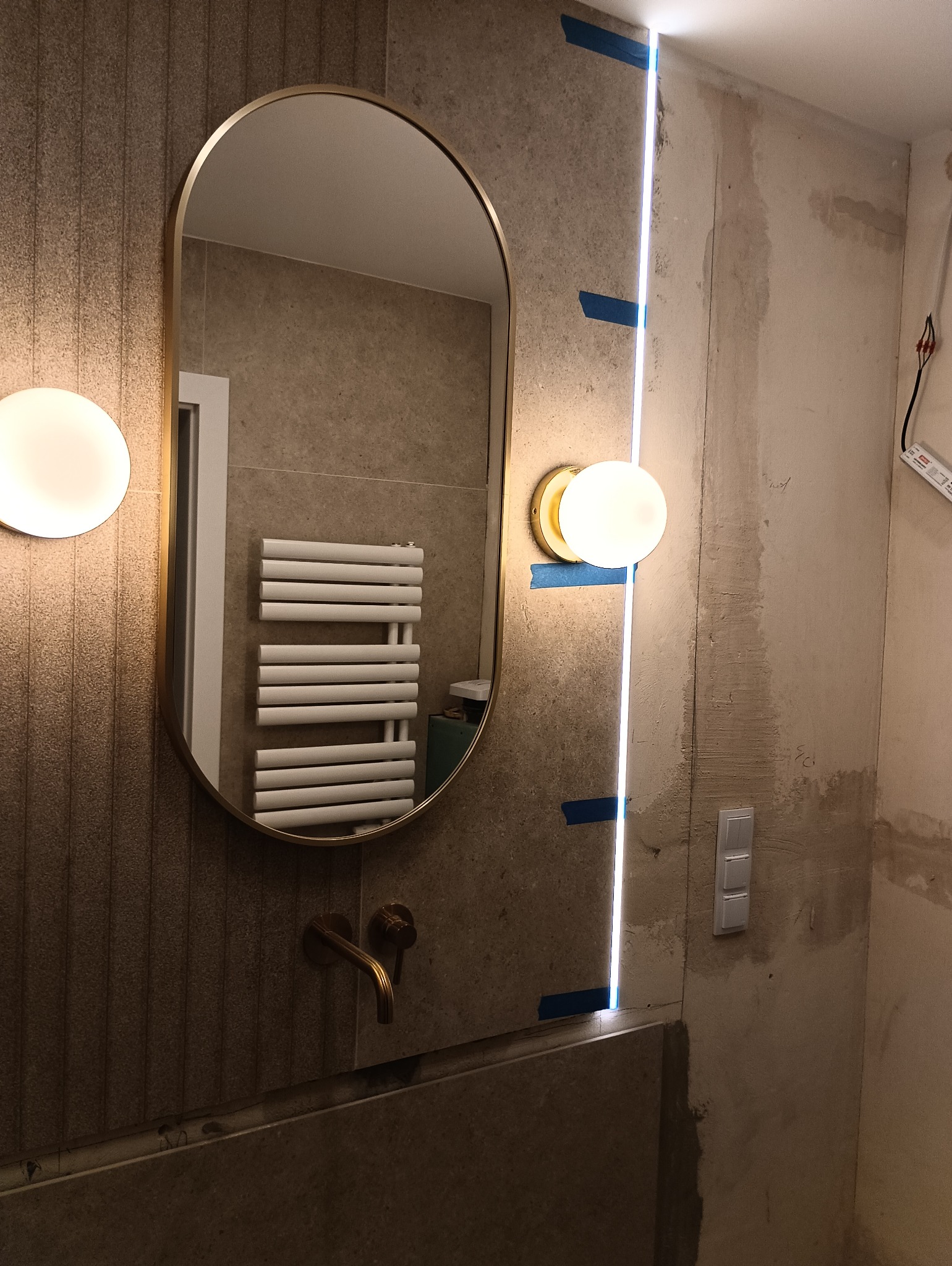 Łazienka w trakcie remontu: owalne lustro w złotej ramie, oświetlenie LED w pionowej szczelinie, widoczne taśmy malarskie, fragment grzejnika drabinkowego w odbiciu lustra, surowe ściany przed...