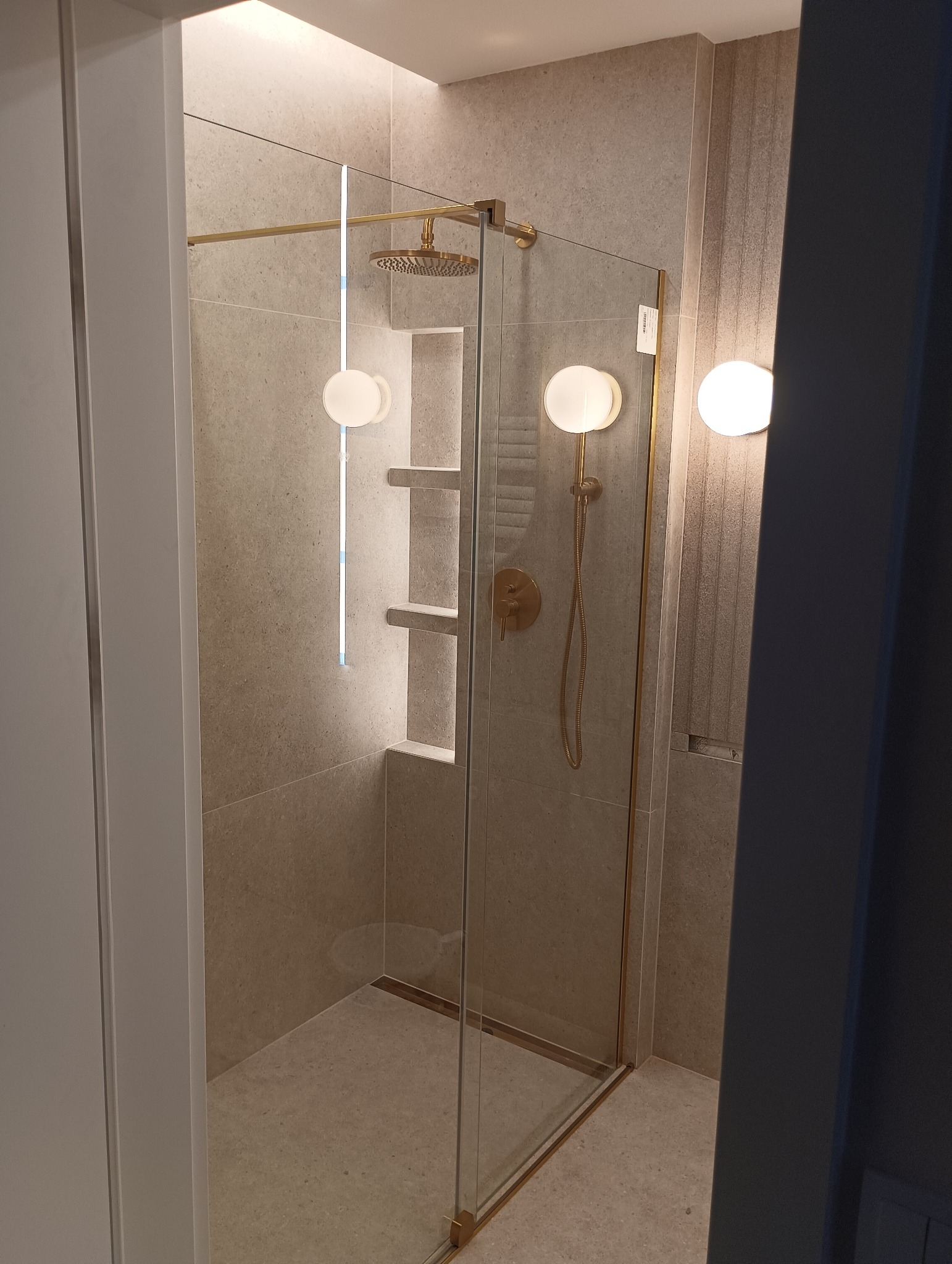 Nowoczesna kabina prysznicowa ze złotymi detalami, deszczownicą, podświetlanymi półkami w ścianie i okrągłymi lampami na ścianie wyłożonej płytkami w odcieniach szarości.