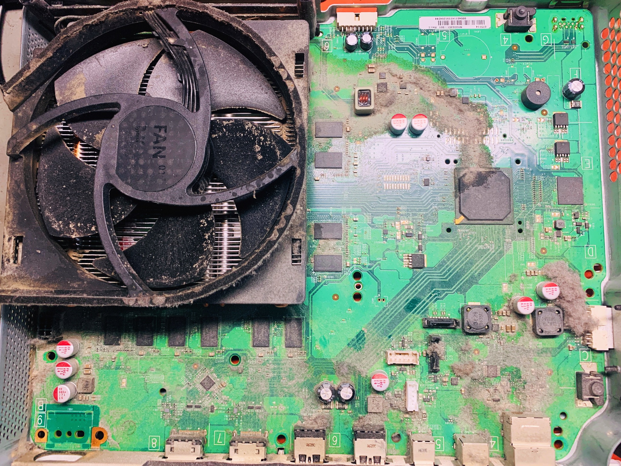 Zabrudzone wnętrze konsoli Xbox z widocznym wentylatorem i płytą główną pokrytą kurzem. Widoczne elementy elektroniczne i złącza. Stan przed czyszczeniem lub naprawą.