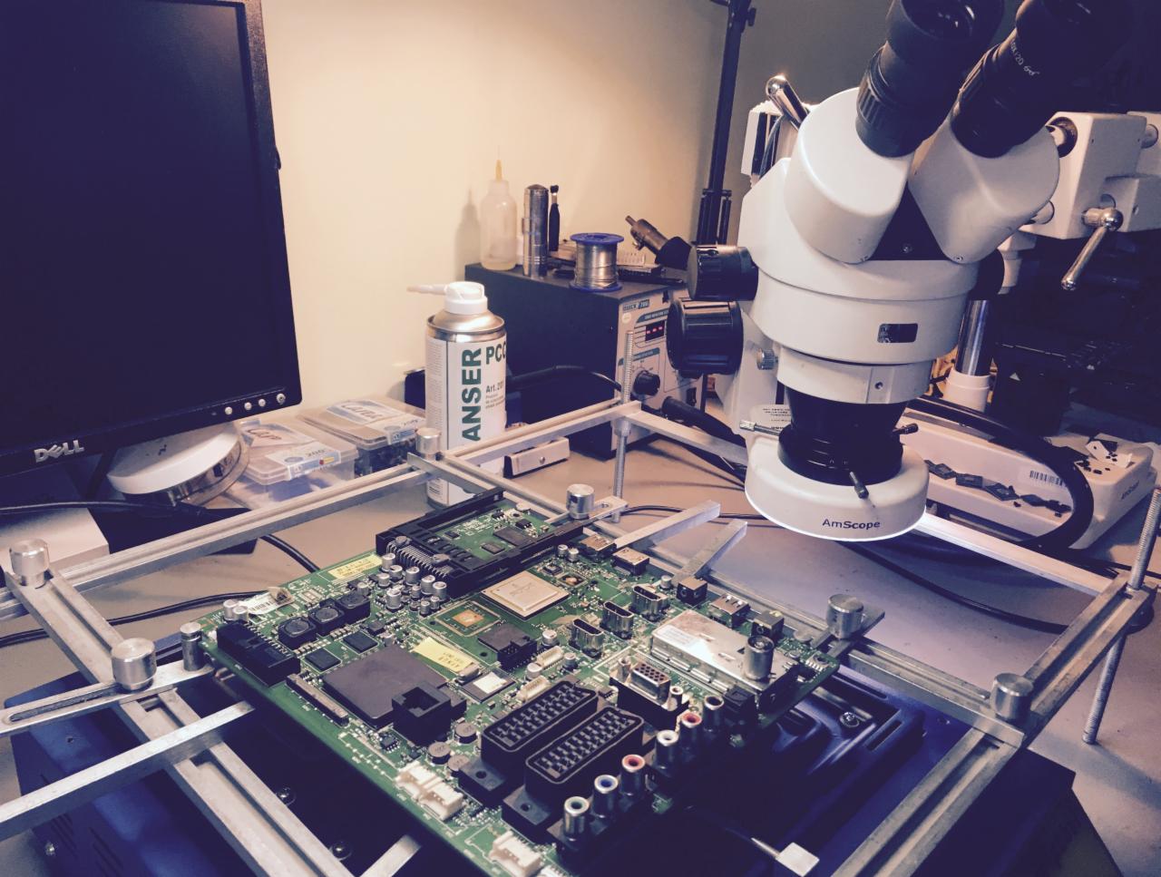 Płyta główna pod mikroskopem AmScope na stanowisku serwisowym, widoczny monitor Dell, puszka ze środkiem czyszczącym Anser PC Art.20 i narzędzia do lutowania