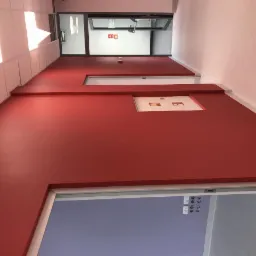 Długi korytarz biurowy z pomalowanymi na czerwono ścianami, białym sufitem z wbudowanym oświetleniem i białymi drzwiami przeciwpożarowymi.