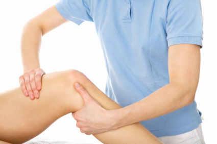 Masaż kolana wykonywany przez osobę w niebieskiej koszulce na białym tle.