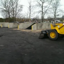 Żółta ładowarka kołowa na placu składowania węgla z betonowymi boksami wypełnionymi czarnym materiałem opałowym, widoczne ślady opon na ziemi.