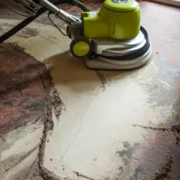 Szlifowanie starej, popękanej posadzki maszyną z zieloną obudową, widoczne ślady po szlifowaniu i pozostałości materiału.