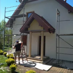 Pracownik na drabinie maluje elewację domu z użyciem wałka, widoczne rusztowania i zabezpieczone podłoże.