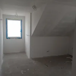 Pokój na poddaszu w trakcie prac wykończeniowych, ściany pokryte świeżą warstwą gładzi, okno zabezpieczone folią malarską, wystające przewody elektryczne.