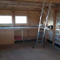 AgMil Usługi remontowo - budowlane - Wnętrze drewnianej altany w trakcie budowy z widoczną konstrukcją ścian z płyty OSB, roboczym stołem z dwoma oknami, drabiną i narzędziami.