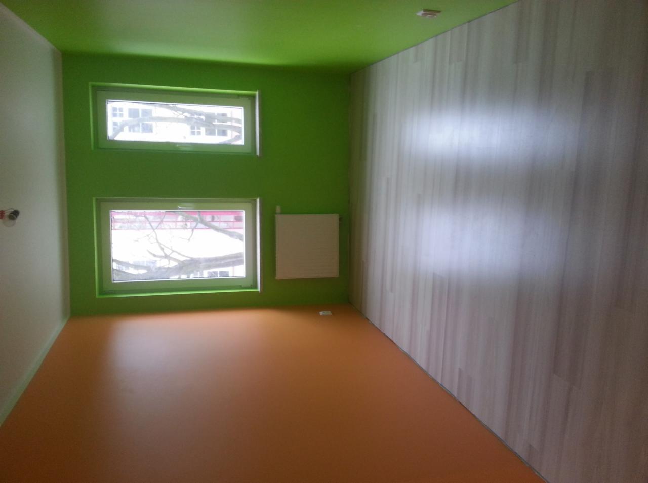 Wnętrze pokoju po remoncie: pomarańczowa podłoga, zielone ściany i sufit, okna z widokiem na drzewo, ściana wyłożona panelami imitującymi drewno.