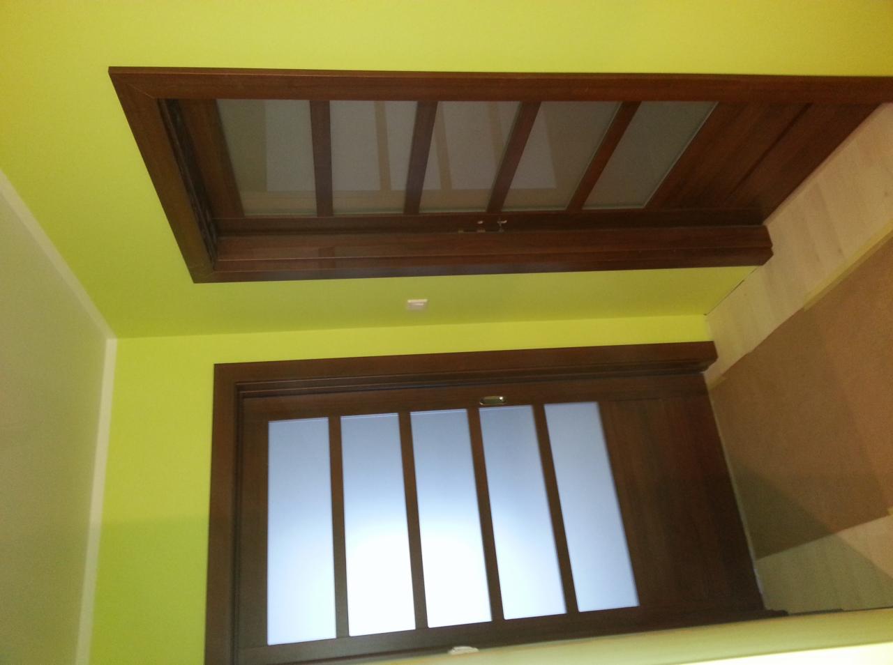 Drewniana konstrukcja antresoli z przeszkleniami, widoczna od dołu, z oświetleniem punktowym na zielonej ścianie.