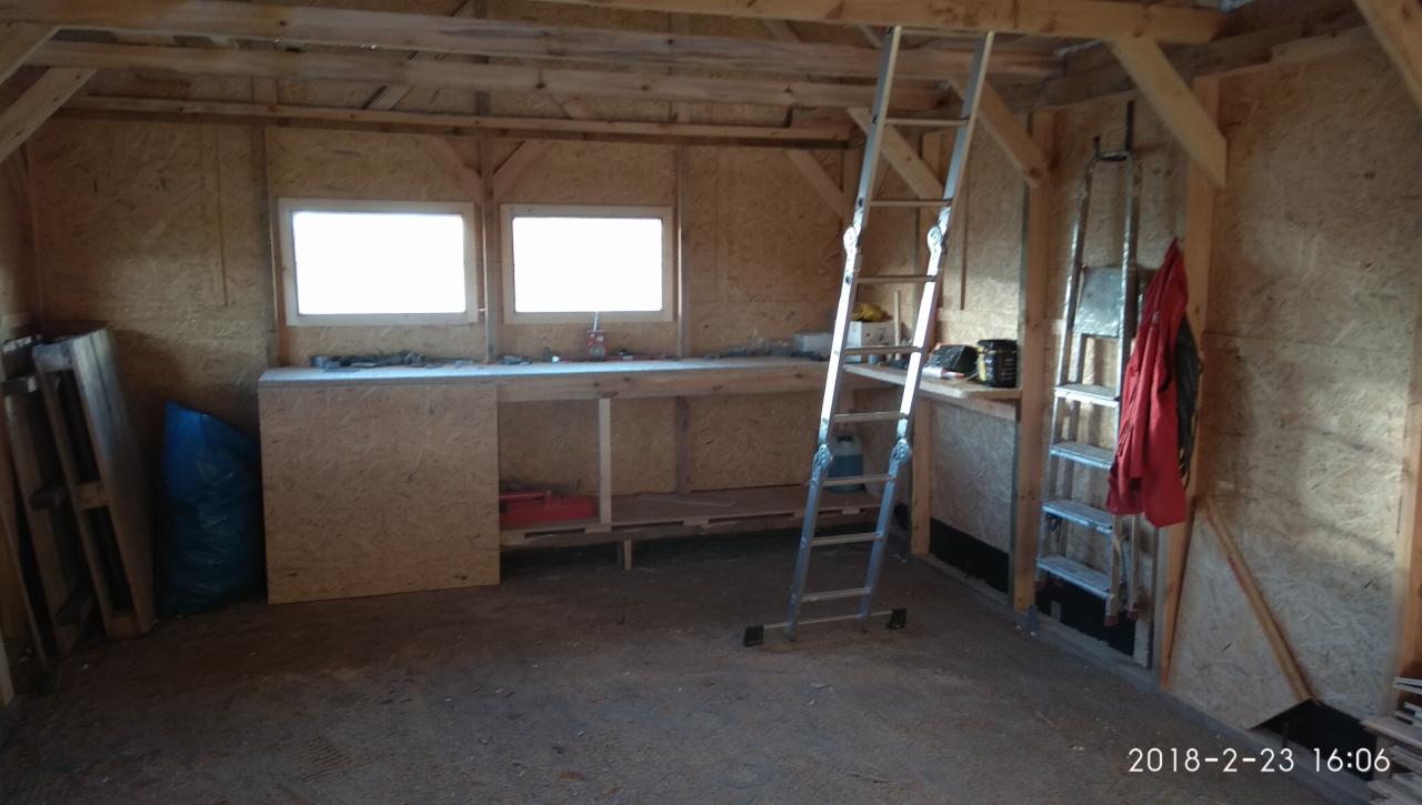 Wnętrze drewnianej altany w trakcie budowy z widoczną konstrukcją ścian z płyty OSB, roboczym stołem z dwoma oknami, drabiną i narzędziami.