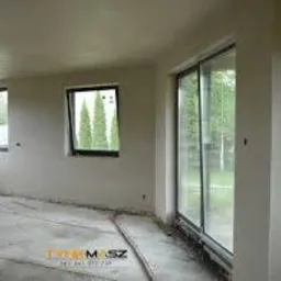 Wnętrze pomieszczenia po nałożeniu tynku maszynowego, widoczne okna PCV, surowa podłoga i ślady po pracach wykończeniowych.