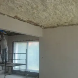 Pracownik w kombinezonie ochronnym na rusztowaniu aplikuje izolację natryskową na sufit wewnątrz budynku w trakcie prac wykończeniowych; okna zabezpieczone folią.