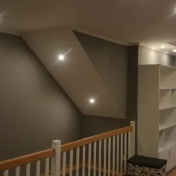 Szare poddasze ze skosem oświetlonym trzema punktowymi lampami LED, biała balustrada schodów, zabudowa okna z szafkami i regałem.