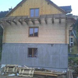 MaxiBud - Surowy stan domu murowanego z drewnianą elewacją w trakcie budowy, widoczne drewniane elementy konstrukcyjne, szara dolna część ściany, palety i deski leżące na ziemi.