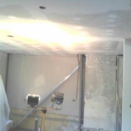 MaxiBud - Wnętrze w trakcie prac wykończeniowych: ściany i sufit pokryte świeżym tynkiem, oświetlone przez halogeny na statywie, widoczne instalacje elektryczne i rury.