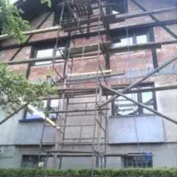 Budynek w trakcie tynkowania elewacji, widoczne rusztowania, surowy mur z cegły oraz fragmenty otynkowanej powierzchni, tło stanowi zieleń drzew.